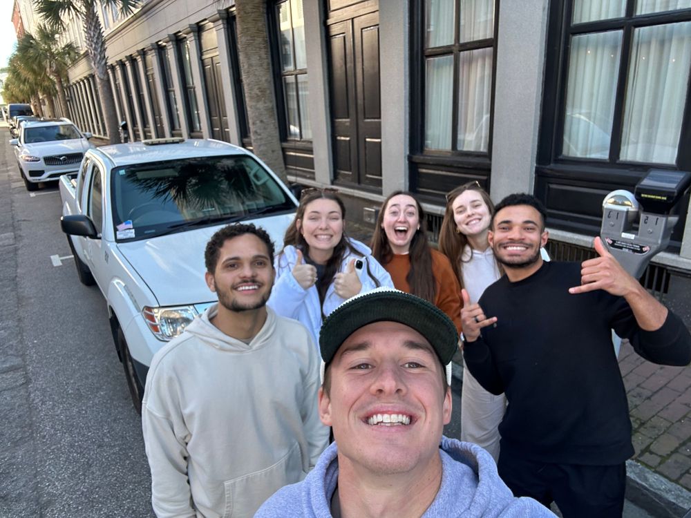 A group of friends taking a street selfie in front of parked cars.