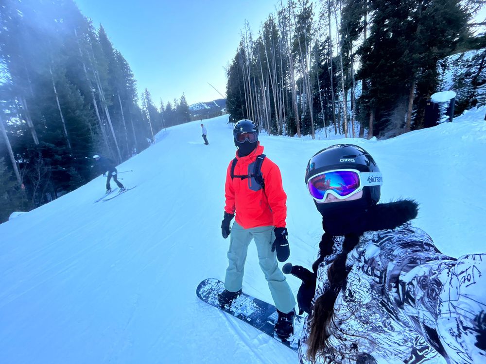Two snowboarders on a snowy ski slope with tall evergreen trees in the background.