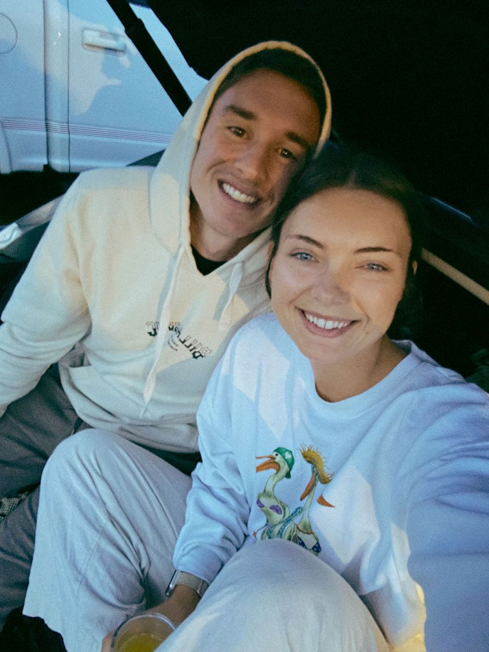 Two people taking a selfie in a car wearing casual sweatshirts.