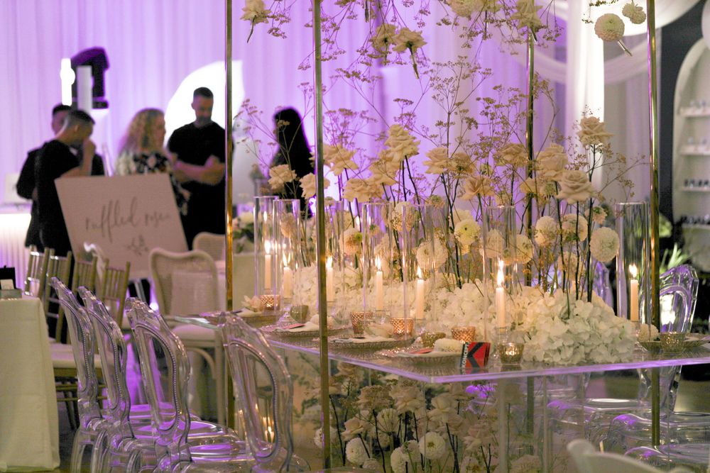 Modern wedding reception featuring glass tables with illuminated floral arrangements and purple accent lighting.
