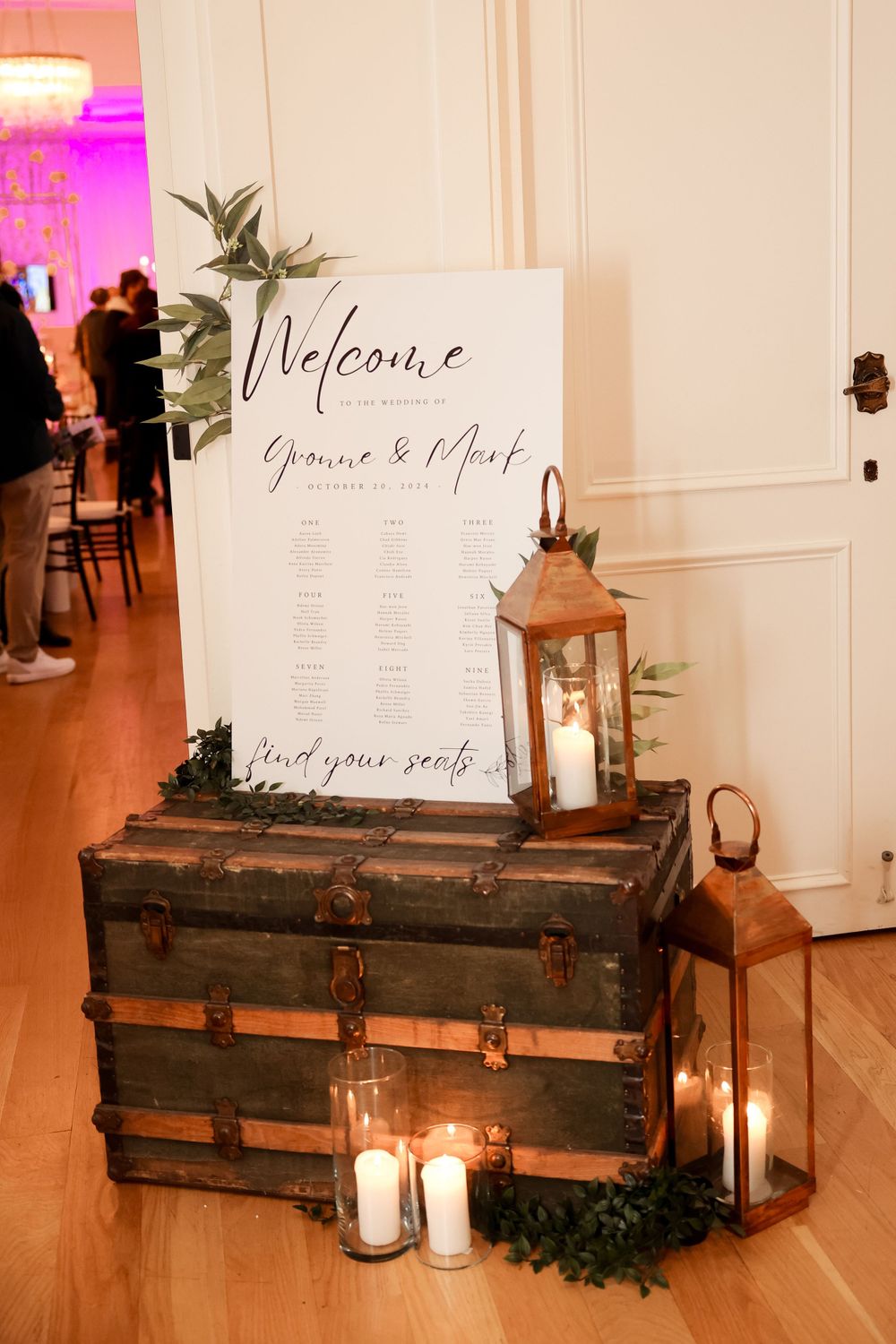 A vintage trunk decorated with lanterns and greenery displays a wedding welcome sign and seating chart.