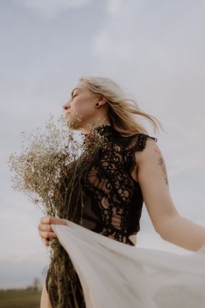 Woman in lace holding dried flowers while her hair flows in the wind against a cloudy sky.