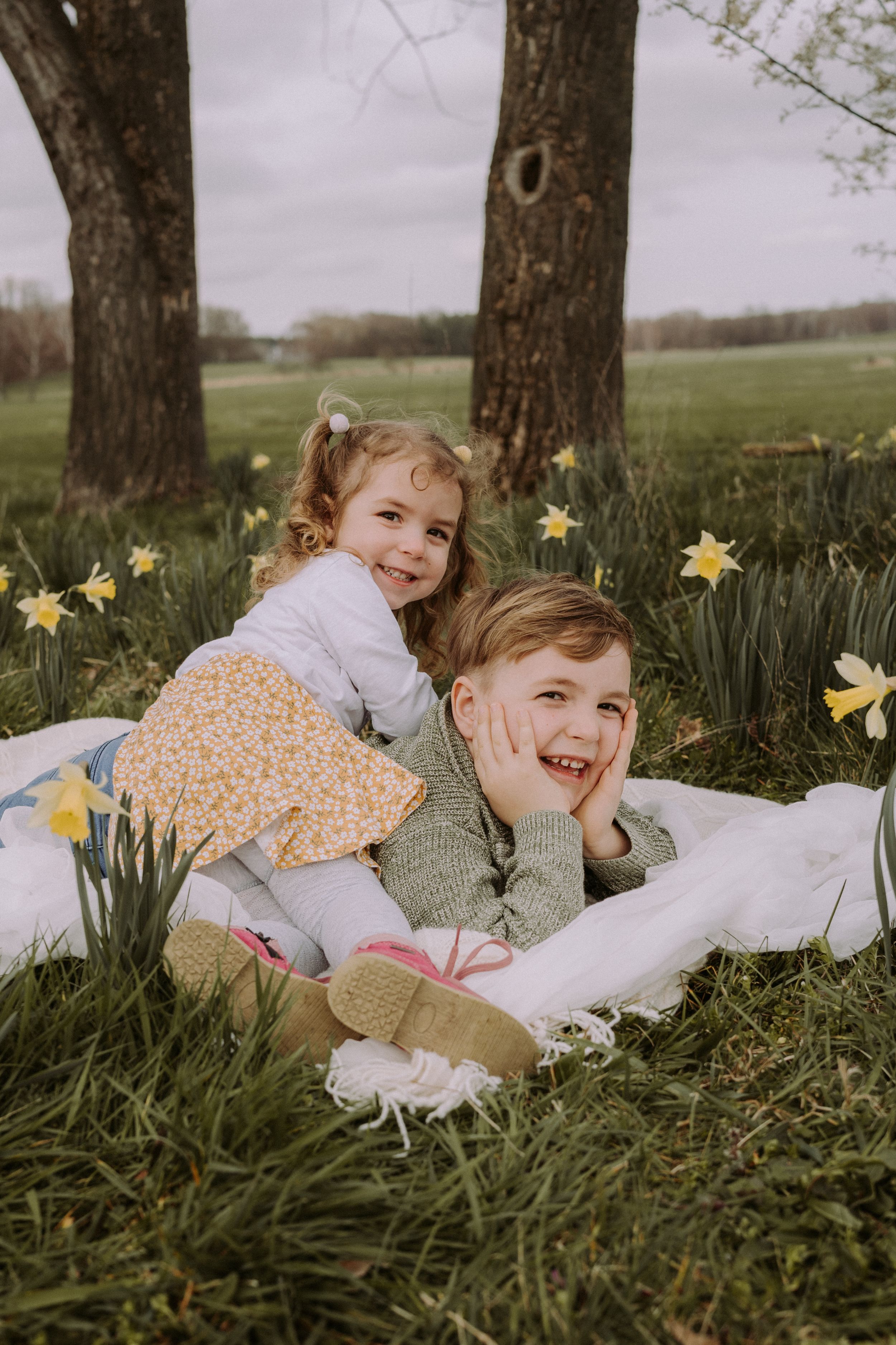 Two children playing and laughing among blooming daffodils under large trees in springtime.