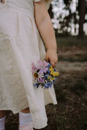 Small hand holding a delicate bouquet of purple, yellow, and blue wildflowers against a white dress.