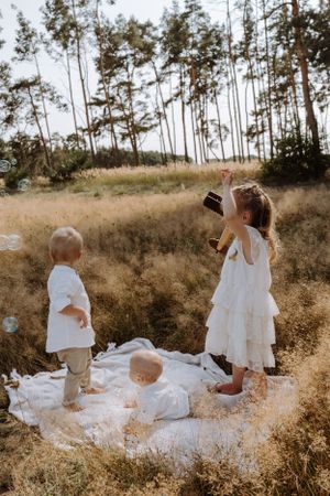 Children playing on a white blanket in a sunny field with tall pine trees in the background.