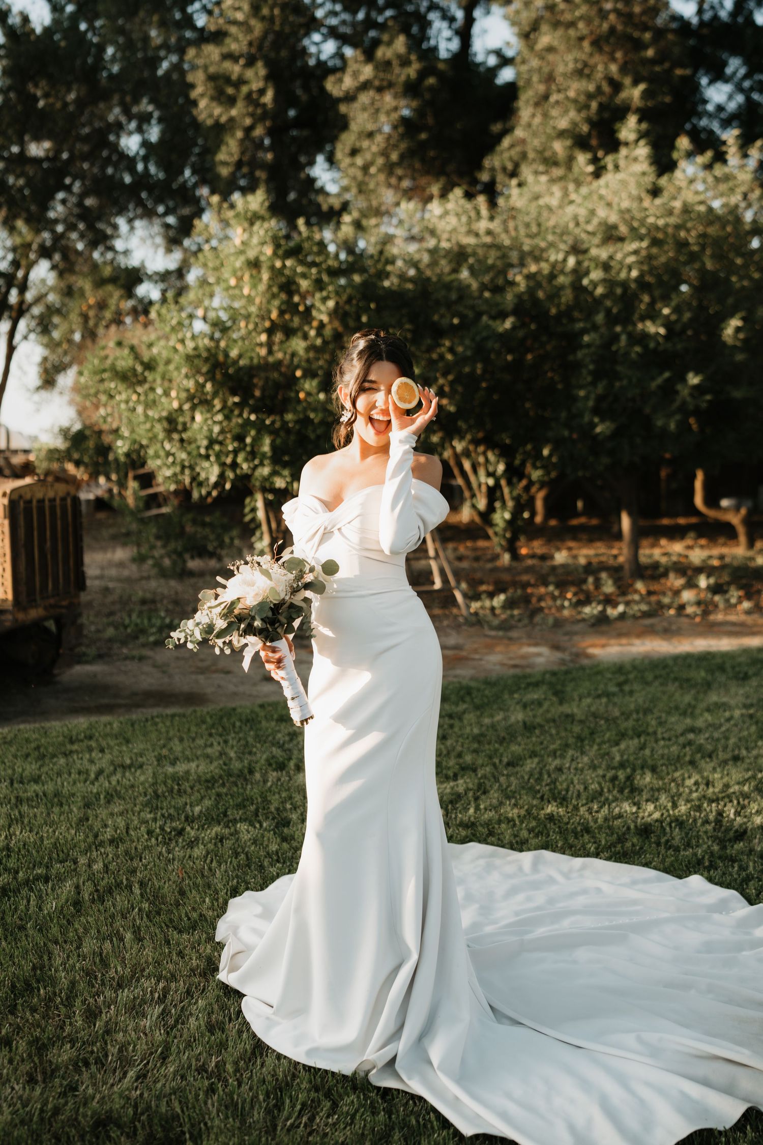 Elegant bride in flowing white dress with ruffled details holds a bouquet during golden hour in a garden setting.