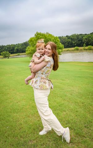 Smiling mother in floral top holding her baby girl on a lush green lawn near a pond.