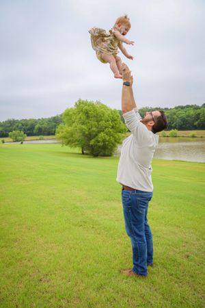 Joyful father tossing baby high in the air on a green lawn with a lake backdrop.