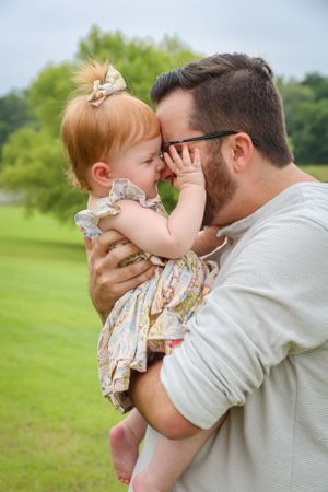 Loving father touching foreheads with his redheaded baby girl in a floral dress outdoors.