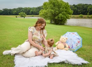 Mother sitting on white blanket with baby daughter and teddy bear on a green lawn.