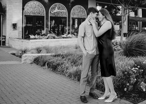 Black and white photo of a couple sharing a romantic kiss on an outdoor plaza with shops in the background.
