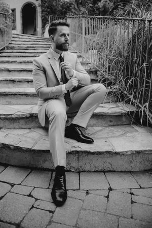 Dapper bearded groom in tan suit sitting on stone steps in black and white photo.