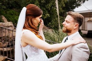 Emotional bride wiping tears while groom smiles tenderly during first look.
