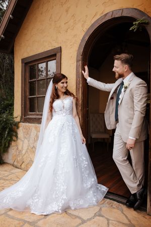 Bride in lace gown and groom in tan suit posing in rustic arched doorway.