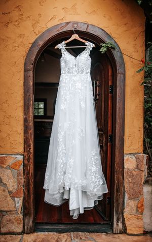White lace wedding dress hanging on wooden hanger in rustic arched doorway.