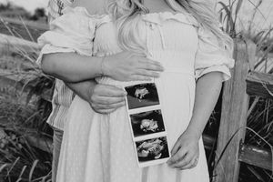 Black and white photo of pregnant couple holding ultrasound photos near rustic fence.