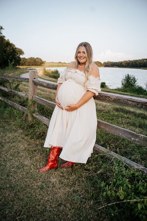 Smiling pregnant woman in white off-shoulder dress and red cowboy boots by lakeside fence.