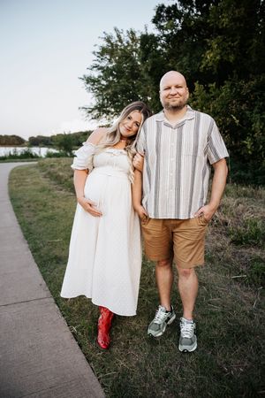 Expecting couple posing together outdoors, woman in white maternity dress with red boots and man in striped shirt.