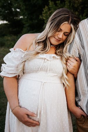 Pregnant woman in white off-shoulder dress cradling baby bump outdoors during golden hour maternity photo session.