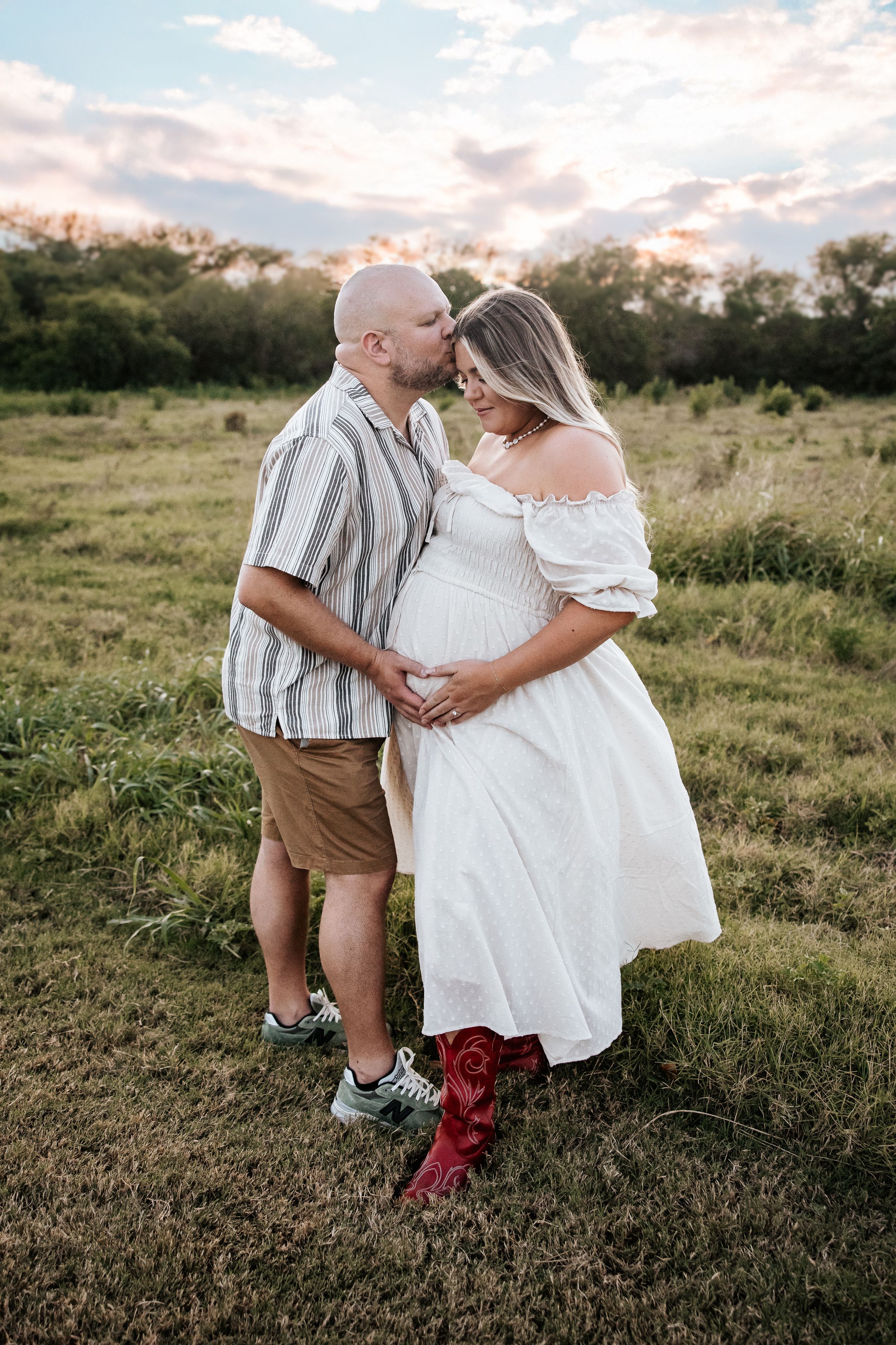 Couple sharing a kiss in green field during sunset maternity photoshoot, hands cradling pregnant belly.