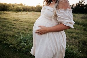 Close-up of pregnant woman in white off-shoulder dress gently holding baby bump in sunlit green field.