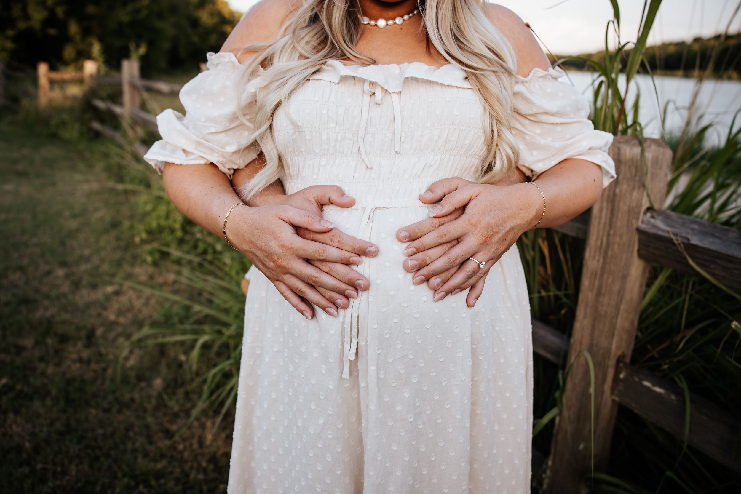 Close-up of pregnant woman in white off-shoulder dress with partner's hands cradling baby bump outdoors at golden hour.