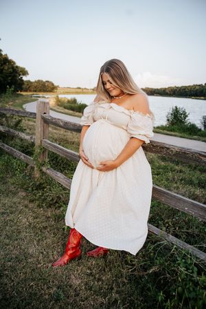 Pregnant woman in white off-shoulder dress and red cowboy boots standing by wooden fence near a lake at sunset.