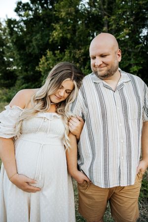Expectant couple standing together outdoors, woman in white maternity dress leaning on partner's shoulder at dusk.