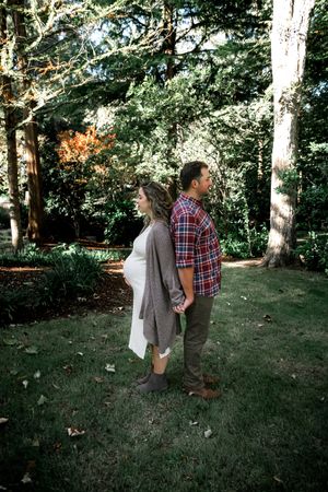 Pregnant couple holding hands back-to-back in a lush green garden park setting during maternity photoshoot.