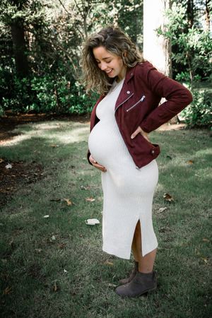 Smiling pregnant woman in white dress and burgundy leather jacket holding her baby bump in a garden.