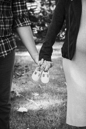 Black and white photo of couple holding hands with tiny baby shoes between them outdoors.