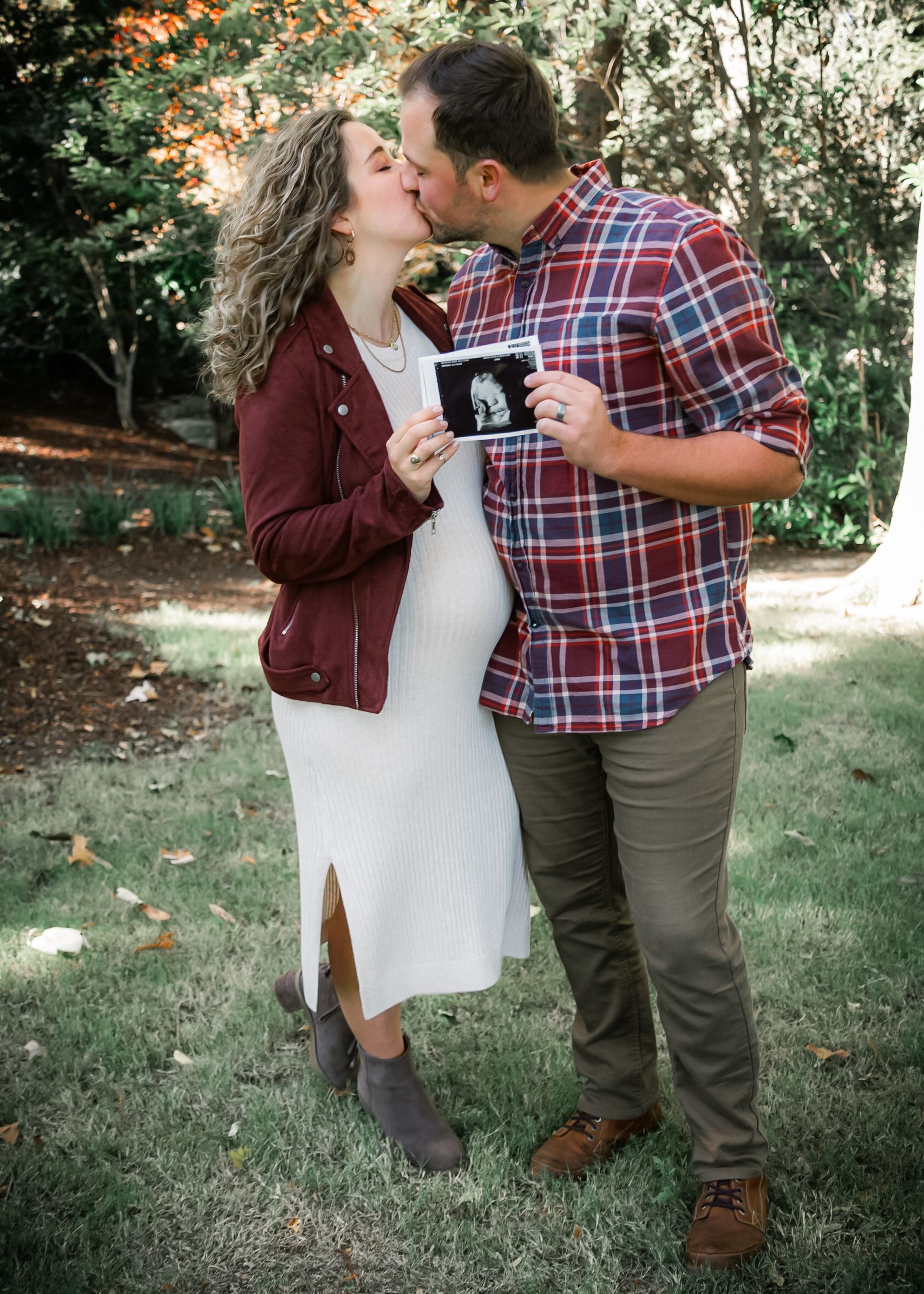 Couple kissing while holding an ultrasound photo during outdoor maternity photoshoot in autumn garden.