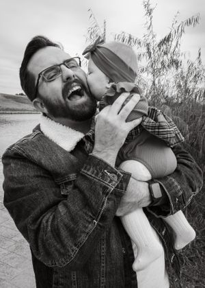 Black and white photo of laughing father being kissed on cheek by his baby near water.