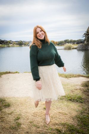Smiling redhead woman in green sweater and white tulle skirt posing by a lakeside.
