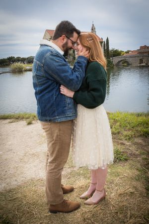 Couple in casual fall outfits touching foreheads romantically beside a scenic lake with bridge.