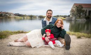 Happy family of three with redhead mother and baby in red hat sitting by a lake.