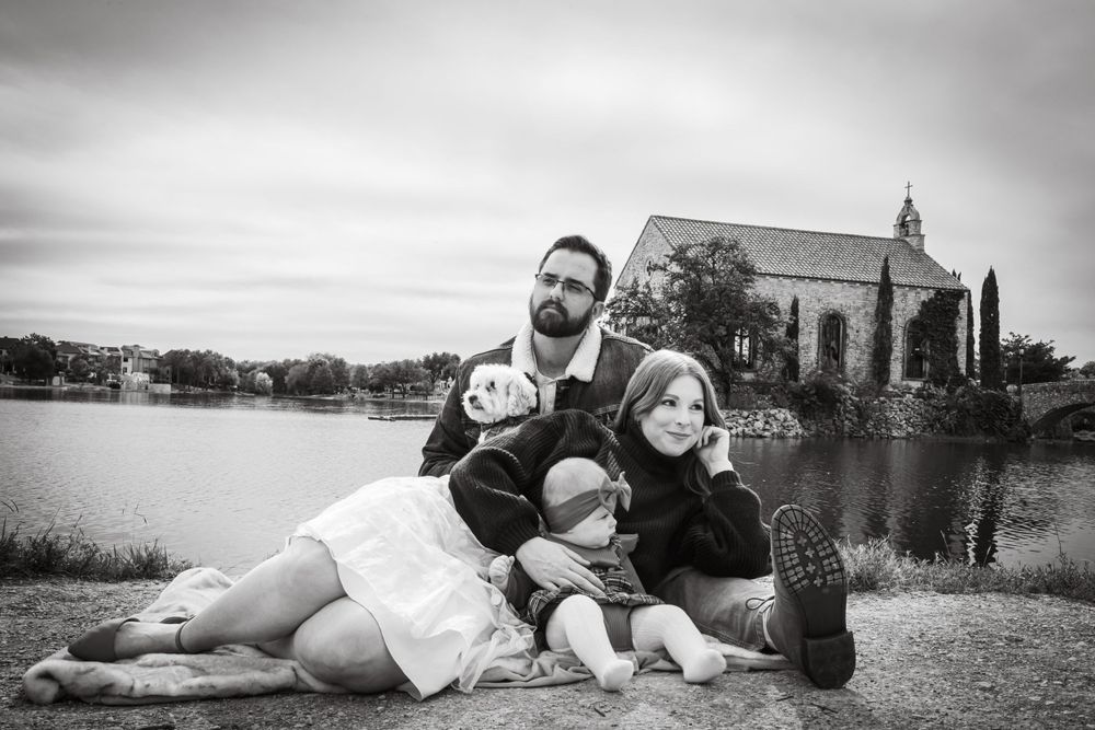 Black and white family portrait with parents and two children sitting by a lake with a chapel in the background.
