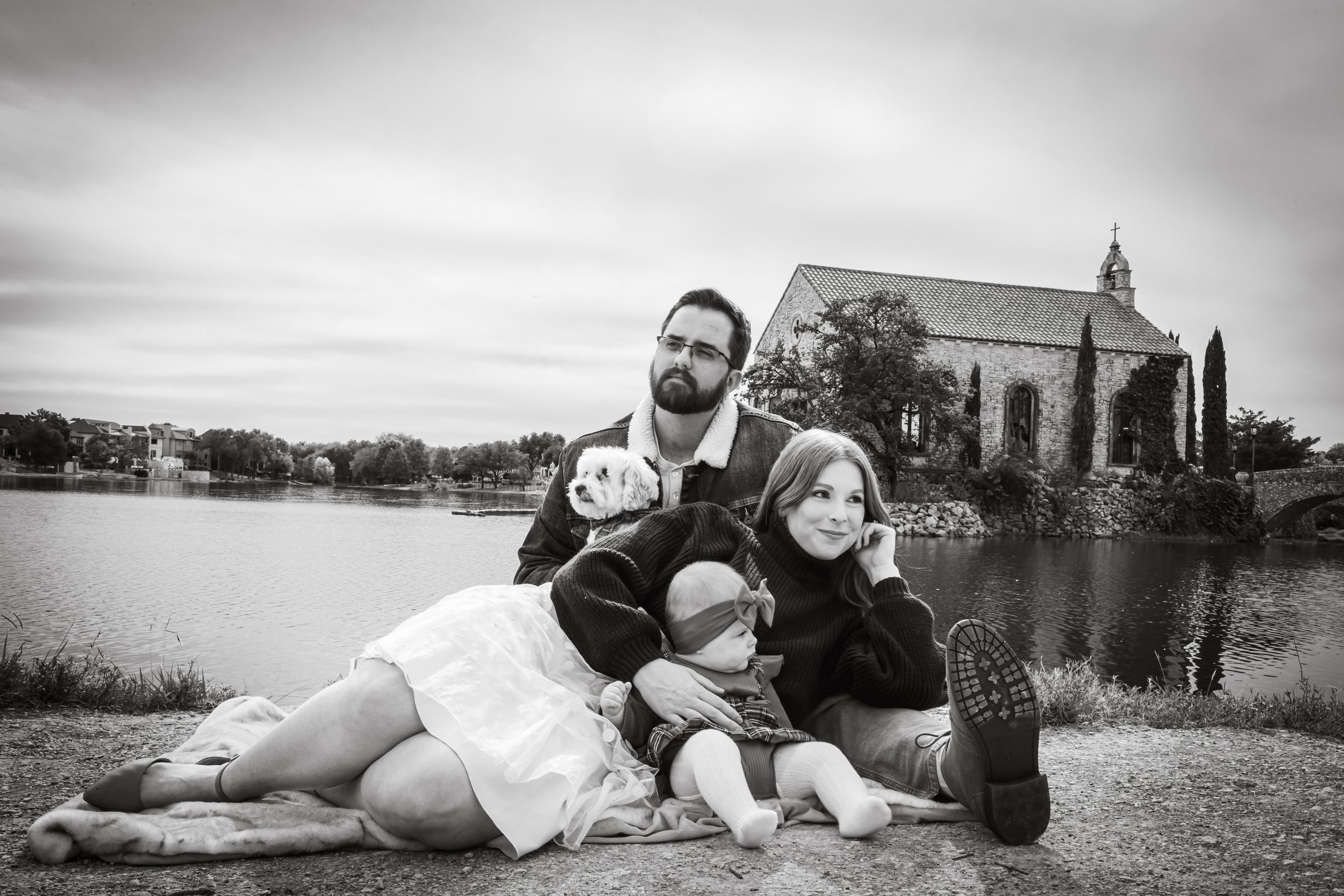 Black and white family portrait with parents and two children sitting by a lake with a chapel in the background.