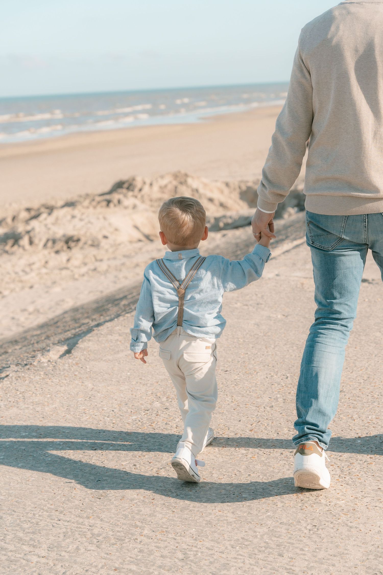 Small child and adult walking hand in hand along a sunny beach shoreline.