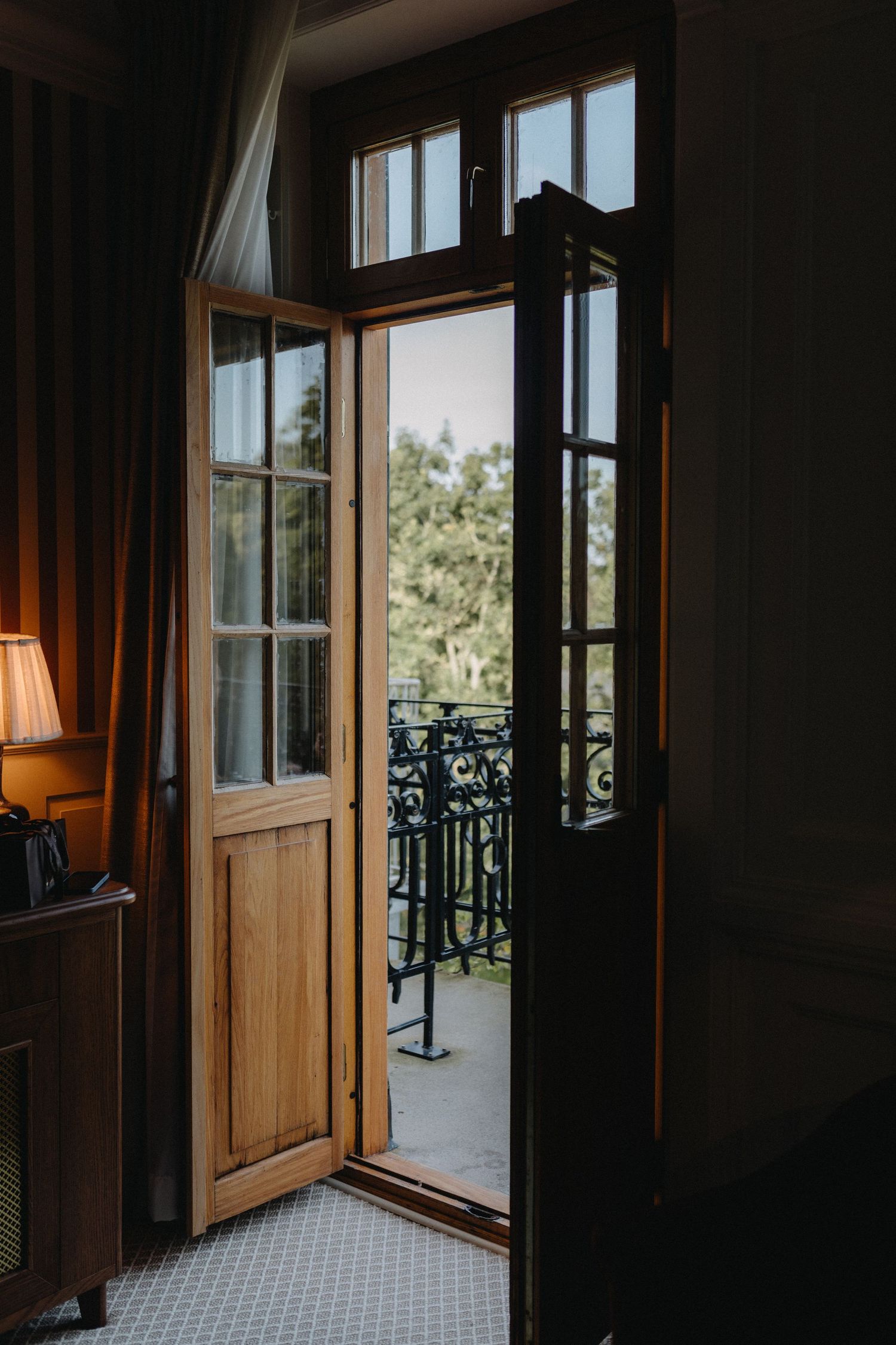 French doors opening to a balcony with ornate iron railings and green foliage views.