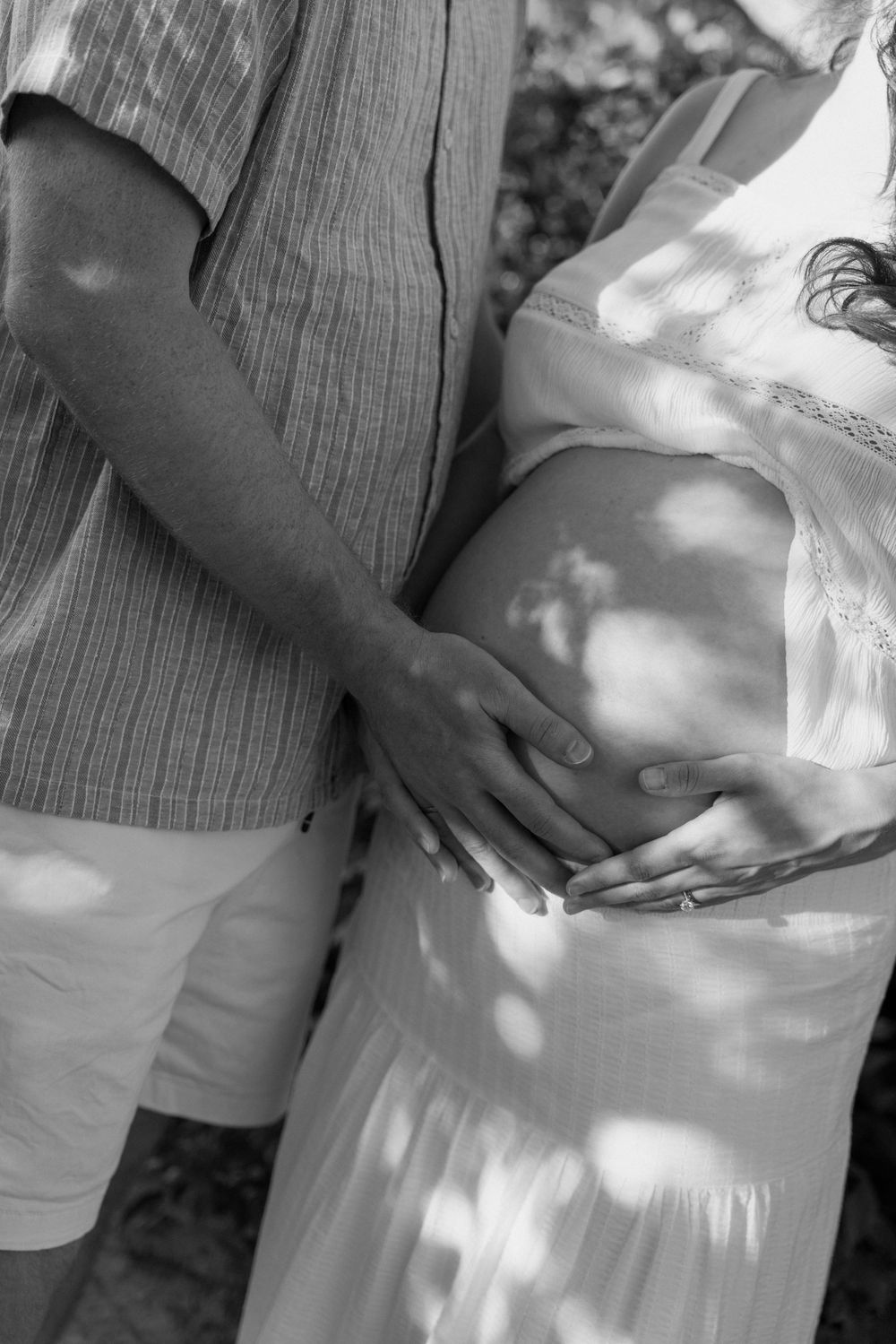Black and white close-up of a couple cradling a pregnant belly with both hands outdoors.