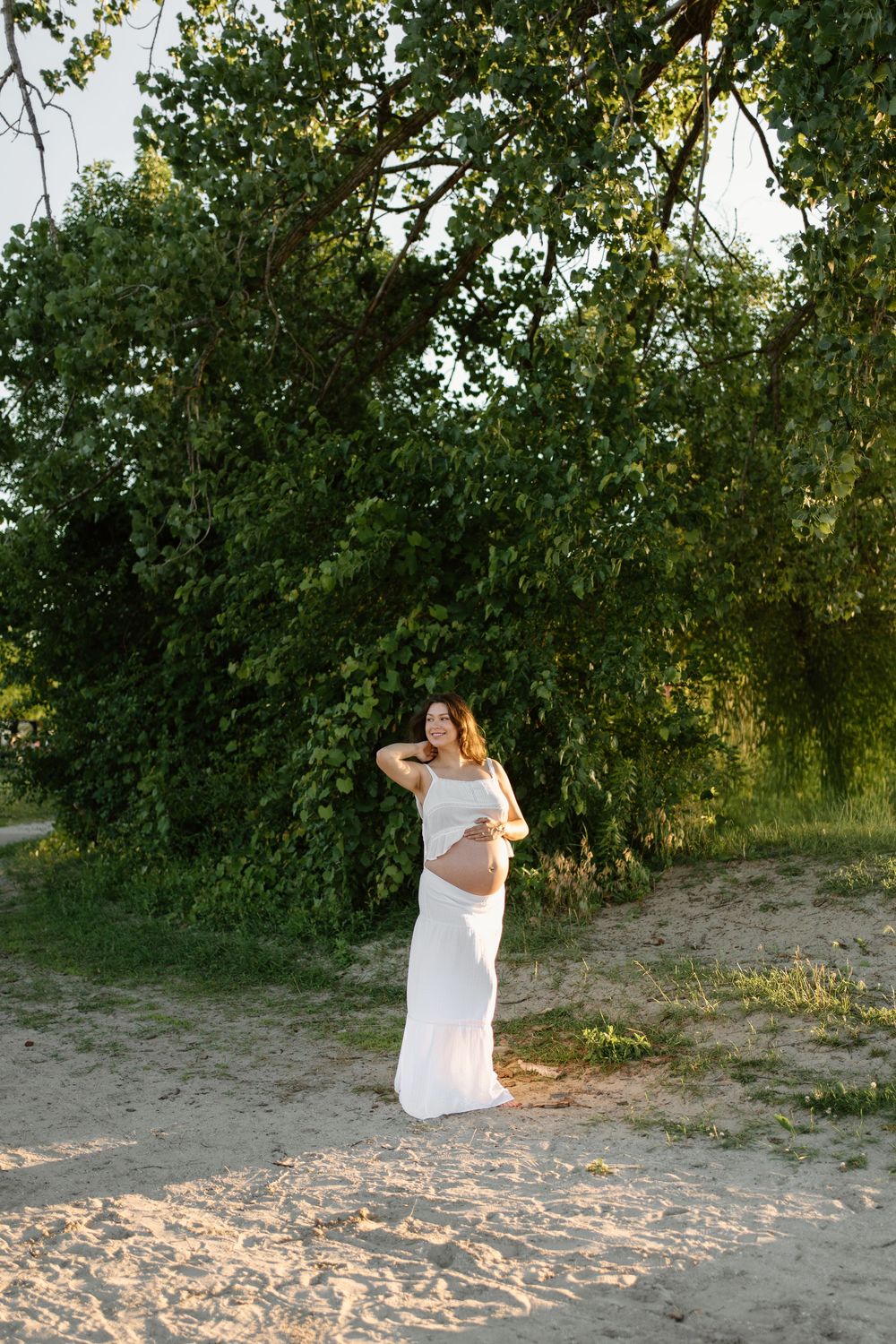 Pregnant woman in a white dress standing on sandy ground surrounded by lush green trees at sunset.