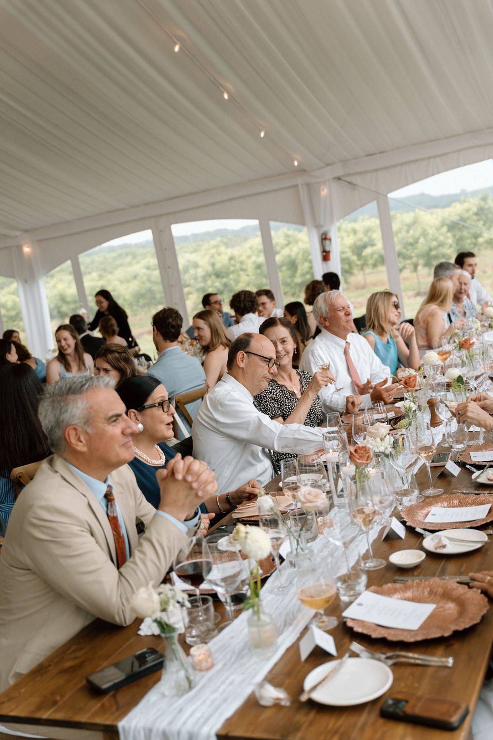 Wedding guests seated at long wooden tables inside a bright tent venue with scenic views during the reception.