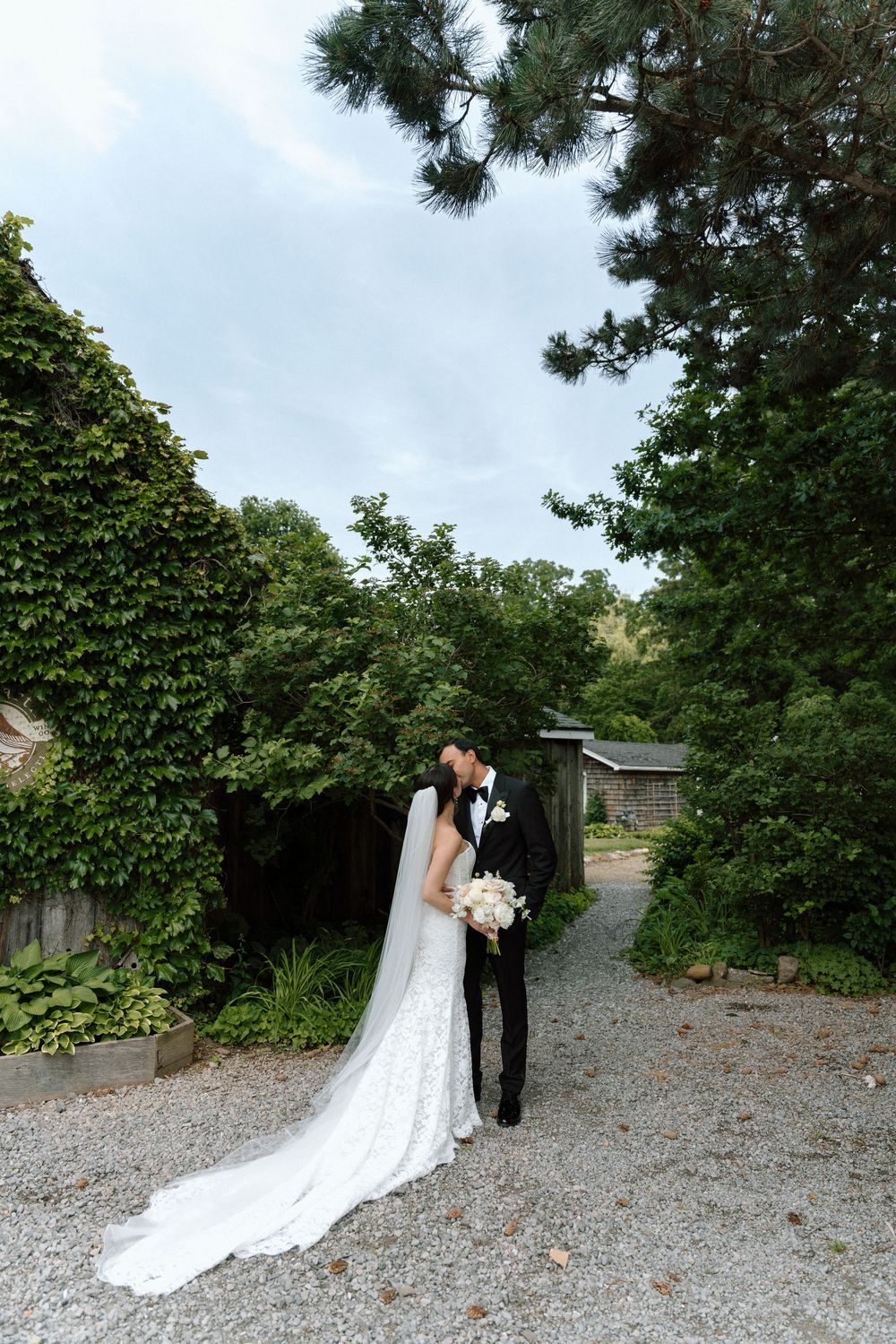 Bride and groom share a kiss on a gravel path surrounded by lush green gardens on their wedding day.