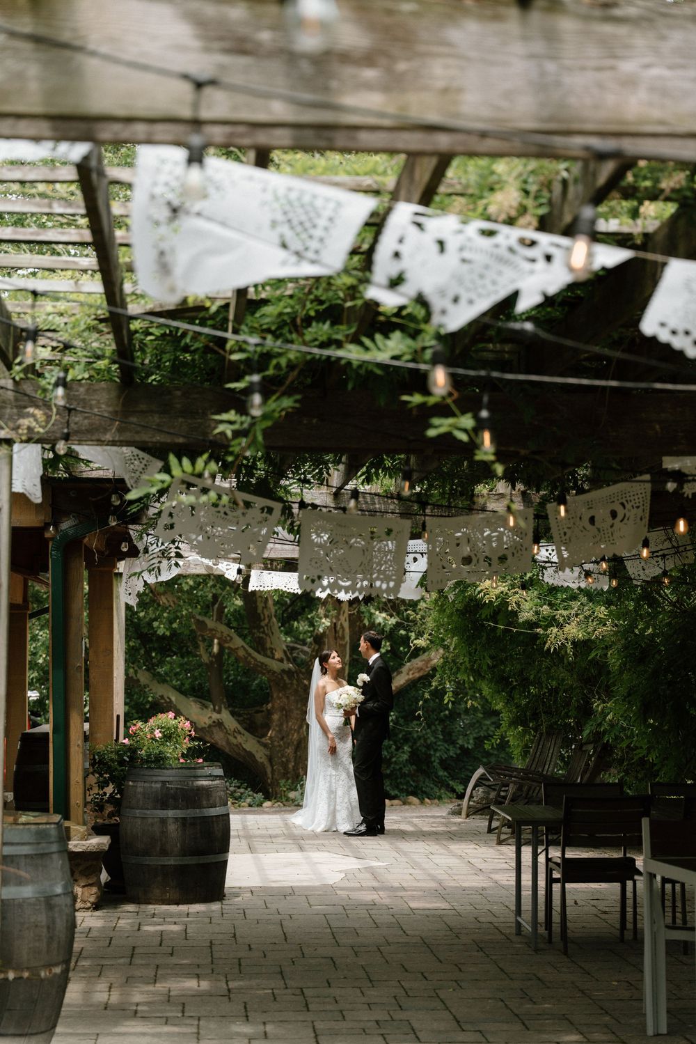 Bride and groom stand together under a rustic pergola adorned with white paper bunting at an outdoor wedding venue.