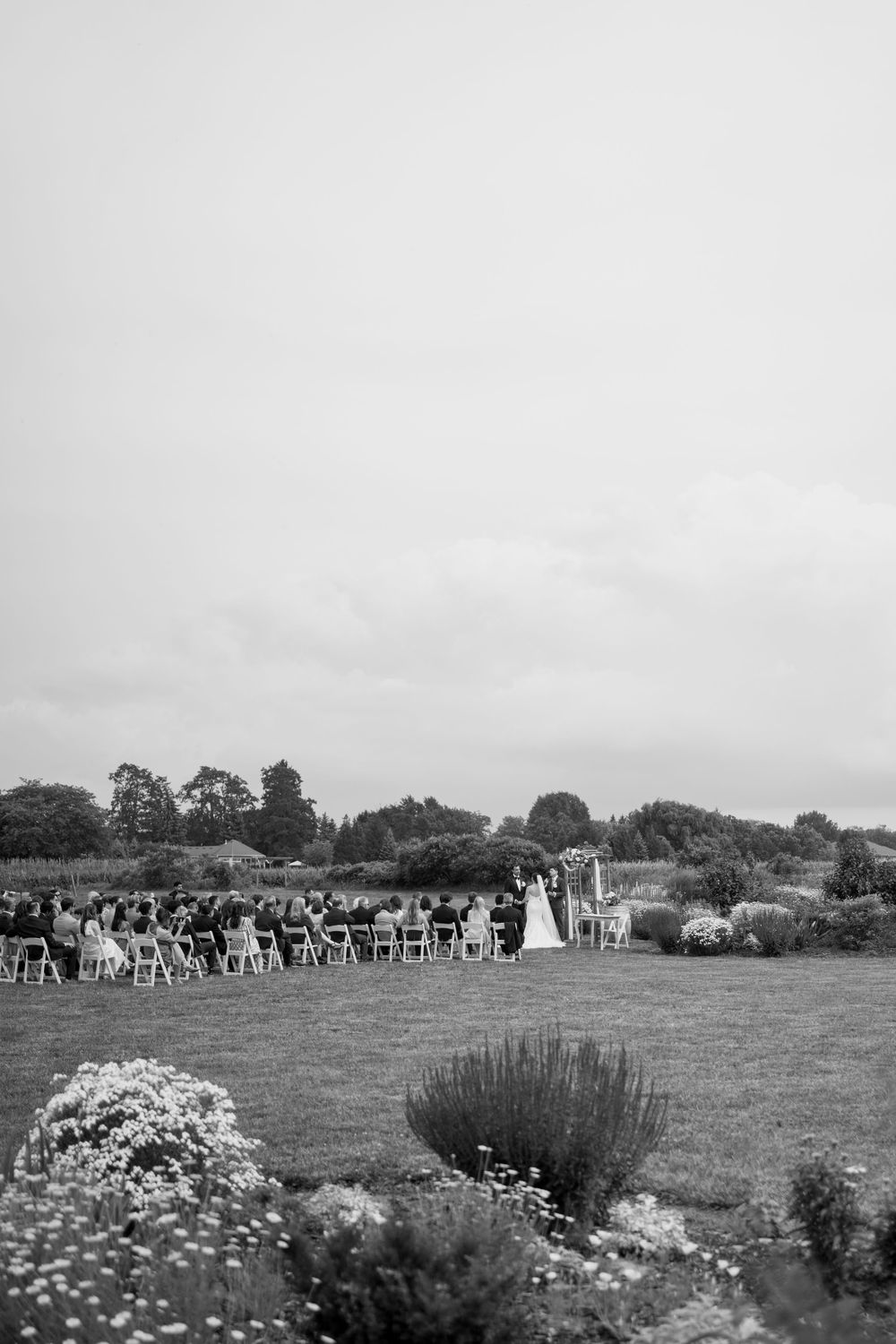 Black and white wide shot of an elegant outdoor wedding ceremony in a lush garden with seated guests.