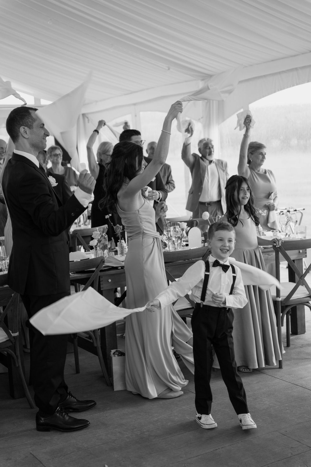 Black and white photo of joyful wedding guests waving napkins as a young boy smiles during the reception.