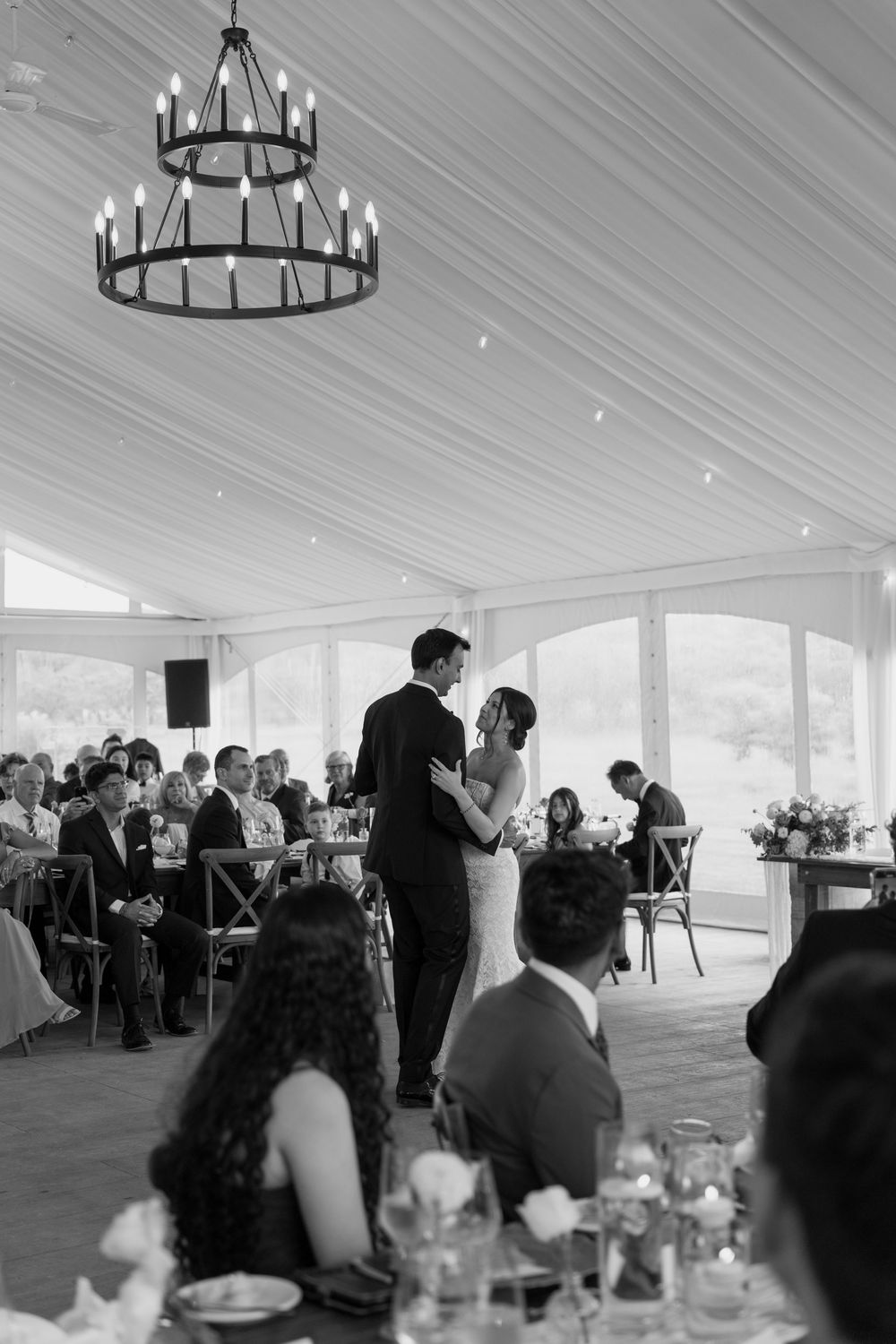 Black and white photo of a bride and groom sharing their first dance under a chandelier at their reception.