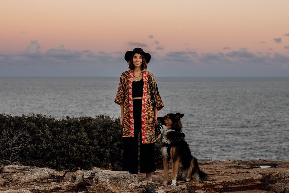 A person in a patterned coat and hat stands with their dog on a rocky coastline in Mallorca at sunset with the ocean in view.