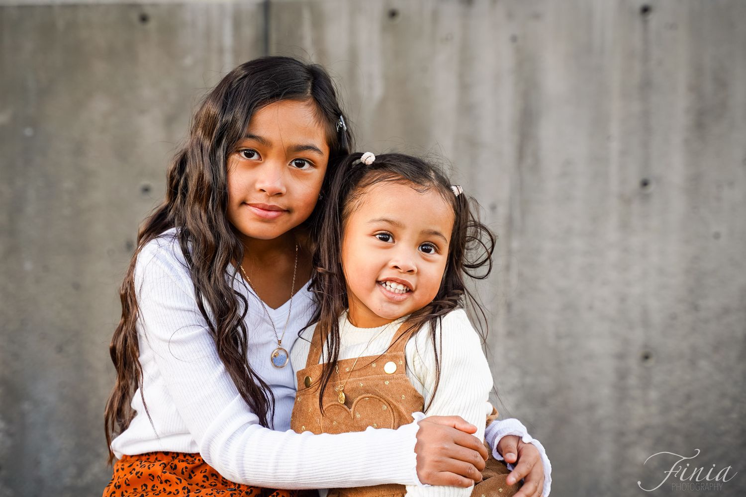 Two children in white tops pose together with a teddy bear against a gray concrete wall.